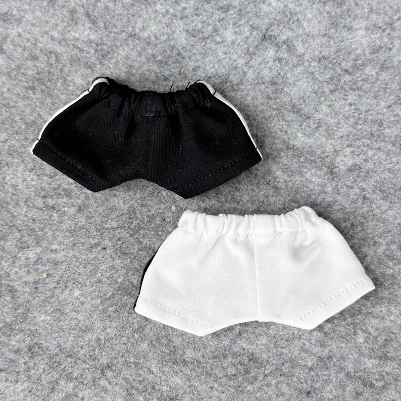 Puede incluir: Dos pares de pantalones cortos deportivos para mu&ntilde;ecas. Uno es negro con rayas laterales blancas, y el otro es blanco con rayas laterales negras. Ambos tienen cinturas el&aacute;sticas y est&aacute;n hechos de tela suave.