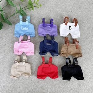 Peut inclure: Ensemble de mini-salopettes de différentes couleurs : bleu avec texte "Lulu", violet, blanc avec texte "B", rose, bleu jean, beige, rouge et noir. Chaque paire a des bretelles réglables et des boucles métalliques.