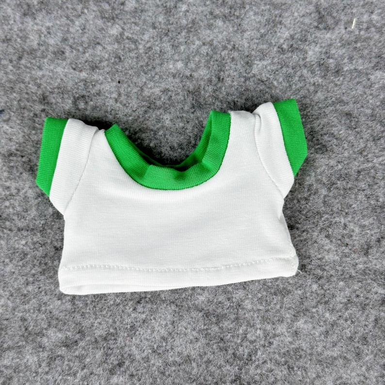 May include: A small white t-shirt with green trim around the collar and sleeves. The shirt is designed for a doll or small toy. The shirt is made of a soft fabric and has a simple, classic design.