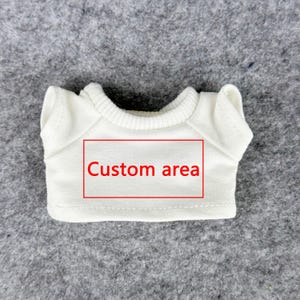 Pode incluir: Uma pequena camiseta branca para boneca, com gola redonda e mangas curtas. A camiseta tem um ret&acirc;ngulo vermelho com a inscri&ccedil;&atilde;o "Custom area" no centro. A camiseta &eacute; feita de material macio e &eacute; exibida em uma superf&iacute;cie cinza.