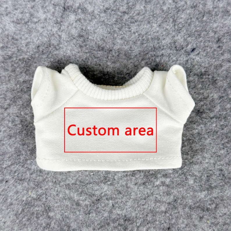 Pode incluir: Uma pequena camiseta branca para boneca, com gola redonda e mangas curtas. A camiseta tem um ret&acirc;ngulo vermelho com a inscri&ccedil;&atilde;o "Custom area" no centro. A camiseta &eacute; feita de material macio e &eacute; exibida em uma superf&iacute;cie cinza.