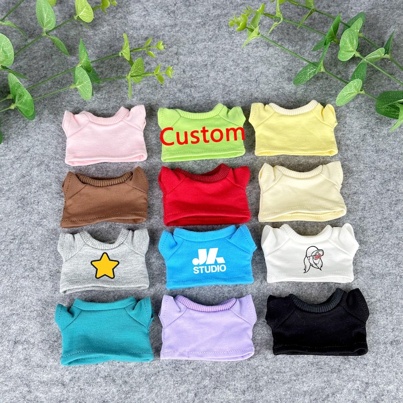 Pode incluir: Uma cole&ccedil;&atilde;o de pequenas camisetas coloridas, cada uma com um design ou texto &uacute;nico. As cores incluem rosa, verde com o texto "Custom", amarelo, castanho, vermelho, creme, cinza com uma estrela, azul com "JL STUDIO", branco com um gr&aacute;fico, turquesa, roxo e preto.