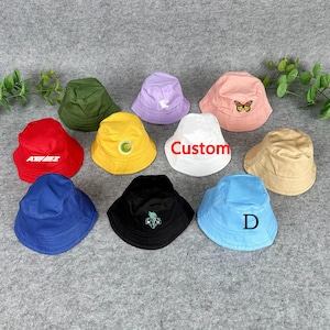 Könnte beinhalten: Eine Sammlung von Fischerhüten in verschiedenen Farben, darunter Rot, Grün, Gelb, Blau, Schwarz und Weiß. Einige Hüte haben gestickte Designs oder Text, wie einen Schmetterling, das Wort "Custom" und die Buchstaben "AE/EZ" und "D".