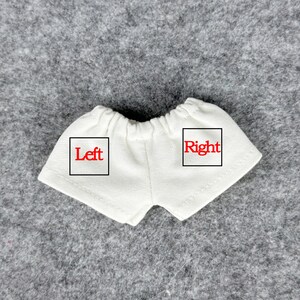Puede incluir: Un par de pantalones cortos blancos para mu&ntilde;ecas con cintura el&aacute;stica. La palabra "Left" est&aacute; impresa en rojo en el lado izquierdo del pantal&oacute;n corto, y la palabra "Right" est&aacute; impresa en rojo en el lado derecho.