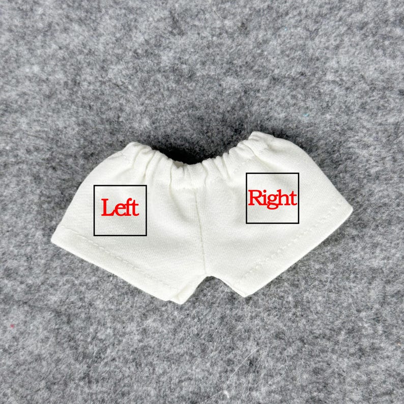 Puede incluir: Un par de pantalones cortos blancos para mu&ntilde;ecas con cintura el&aacute;stica. La palabra "Left" est&aacute; impresa en rojo en el lado izquierdo del pantal&oacute;n corto, y la palabra "Right" est&aacute; impresa en rojo en el lado derecho.