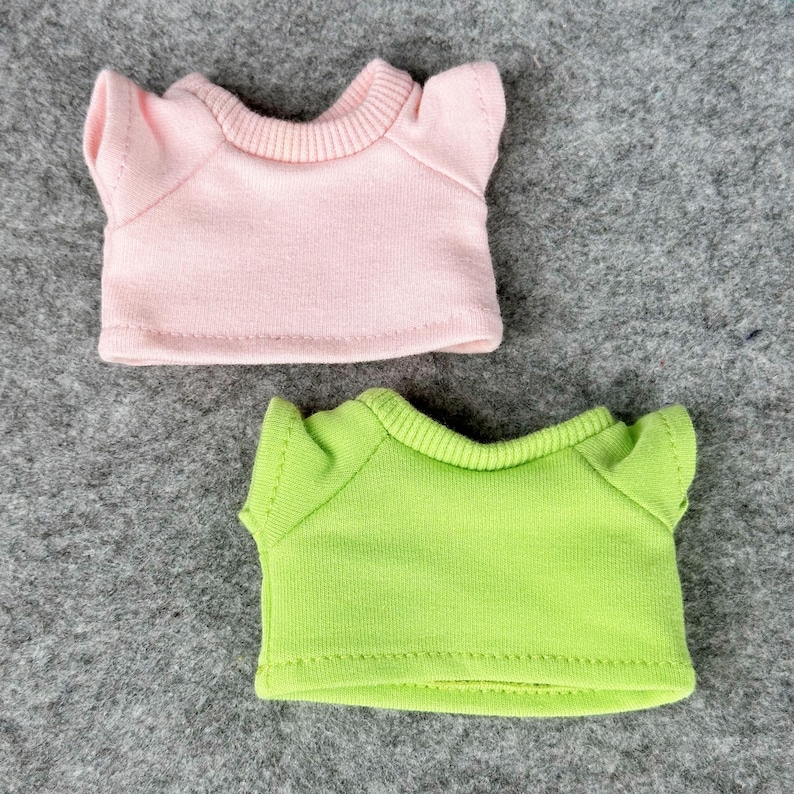 Pode incluir: Duas pequenas camisetas de boneca. Uma &eacute; rosa claro, a outra verde lim&atilde;o. Ambas t&ecirc;m mangas curtas e gola canelada. As camisas s&atilde;o feitas de um tecido macio e s&atilde;o projetadas para bonecas.