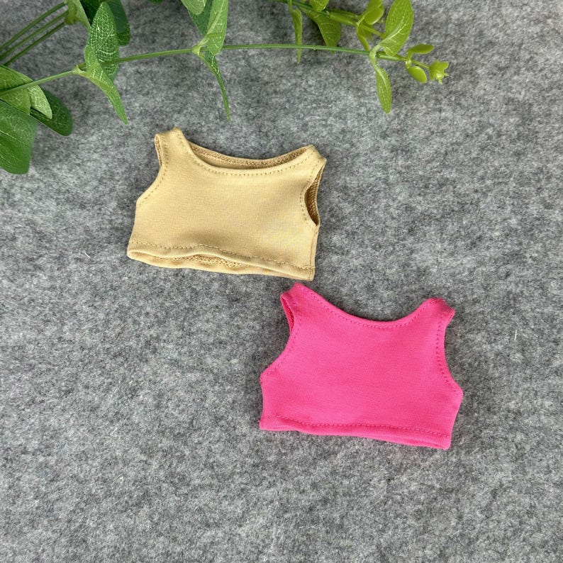 Peut inclure: Deux petits d&eacute;bardeurs, un beige et un rose vif. Les hauts sont faits d'une mati&egrave;re douce et sont con&ccedil;us sans manches. Ils sont probablement destin&eacute;s &agrave; une poup&eacute;e ou &agrave; une petite figurine. Fond gris.