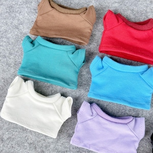 Pode incluir: Uma cole&ccedil;&atilde;o de camisetas de boneca em miniatura em v&aacute;rias cores, incluindo marrom, vermelho, verde-azulado, azul, branco e lavanda. Cada camisa tem gola redonda e mangas curtas, dispostas em uma superf&iacute;cie cinza.