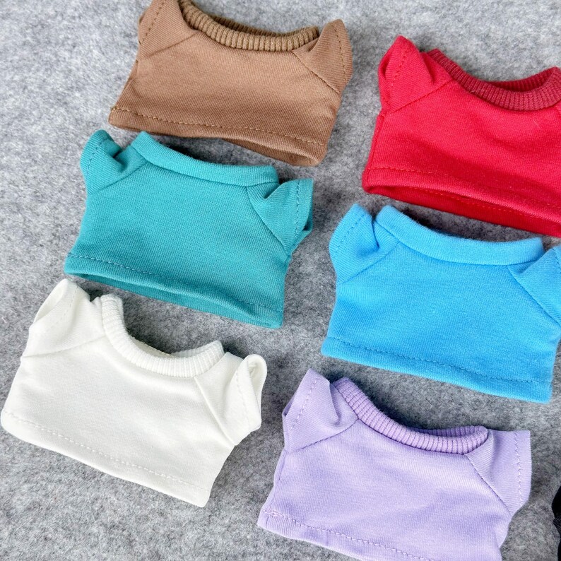 Pode incluir: Uma cole&ccedil;&atilde;o de camisetas de boneca em miniatura em v&aacute;rias cores, incluindo marrom, vermelho, verde-azulado, azul, branco e lavanda. Cada camisa tem gola redonda e mangas curtas, dispostas em uma superf&iacute;cie cinza.