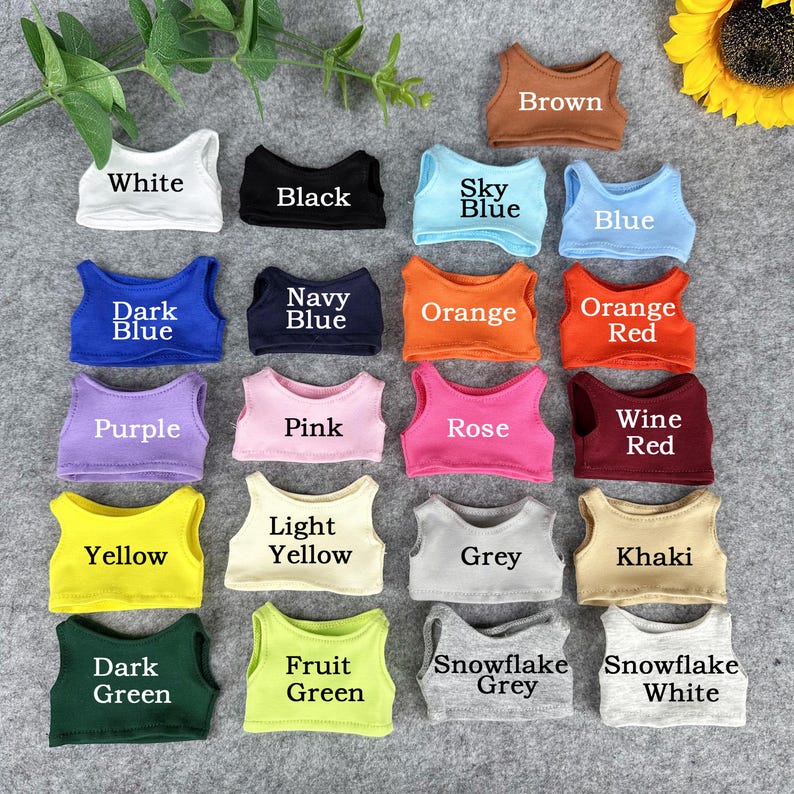 Peut inclure: Une collection de petits d&eacute;bardeurs de poup&eacute;e dans diverses couleurs, dont le blanc, le noir, le bleu et le vert. Chaque haut est &eacute;tiquet&eacute; avec son nom de couleur, comme "Bleu ciel" et "Rouge vin". Les hauts sont dispos&eacute;s sur une surface grise.