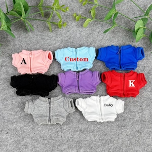May include: A collection of miniature zip-up jackets in various colors, including pink, blue, black, purple, red, gray, and white. Some jackets have text: "A", "Custom", "K", and "Baby". The jackets are designed for dolls or small toys.