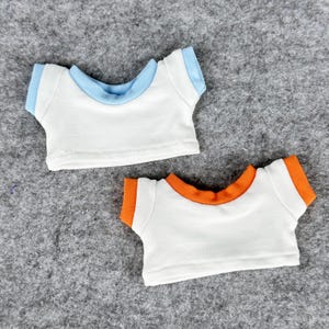 May include: Two small white t-shirts with contrasting trim. One has light blue trim on the collar and sleeves, and the other has orange trim. These doll-sized shirts are designed to fit small plush toys.