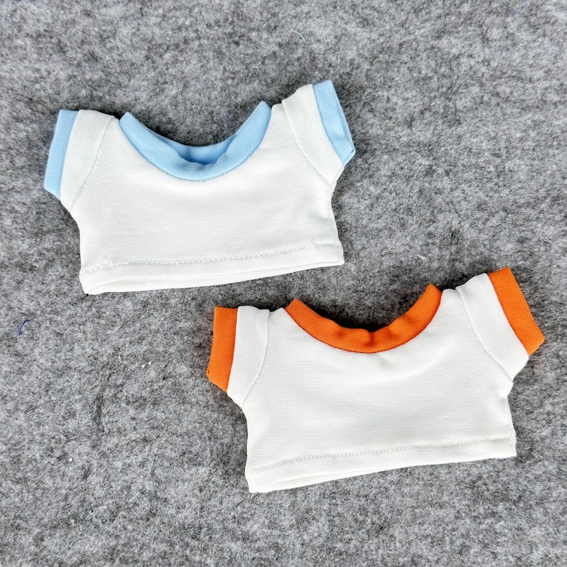 May include: Two small white t-shirts with contrasting trim. One has light blue trim on the collar and sleeves, and the other has orange trim. These doll-sized shirts are designed to fit small plush toys.