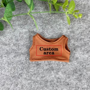 Peut inclure: Un petit haut sans manches marron avec le texte "Custom area" dans un rectangle rouge. Le haut est fait d'un tissu doux et est probablement destin&eacute; &agrave; une poup&eacute;e ou une petite figurine. Des feuilles vertes sont visibles en arri&egrave;re-plan.
