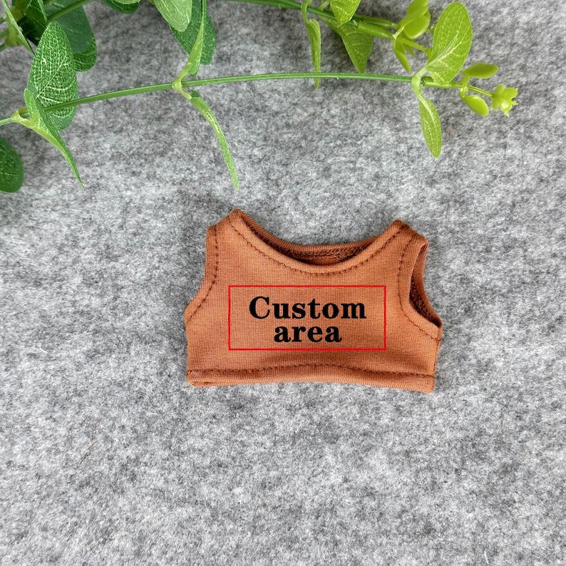 Peut inclure: Un petit haut sans manches marron avec le texte "Custom area" dans un rectangle rouge. Le haut est fait d'un tissu doux et est probablement destin&eacute; &agrave; une poup&eacute;e ou une petite figurine. Des feuilles vertes sont visibles en arri&egrave;re-plan.