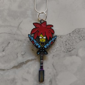 Puede incluir: Collar de cadena plateada con un colgante que representa un personaje de dibujos animados con pelo rojo, cara amarilla y cuchillos azules. El colgante tiene un acabado metálico iridiscente en la parte inferior.