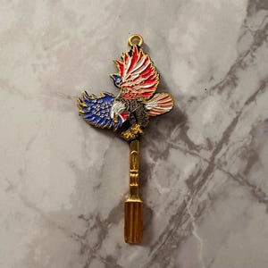 May include: A gold-toned metal spoon featuring an enamelled eagle emblem at the top. The eagle is depicted with wings spread, showcasing a patriotic design with red, white, and blue colours, including white stars on a blue background on one wing.