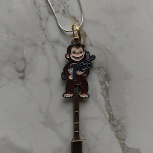 Puede incluir: Collar de cadena plateada con un colgante que representa un mono de dibujos animados sosteniendo un rifle. El mono es marrón con detalles en blanco y negro. El colgante está unido a un dije en forma de cuchara de color bronce.
