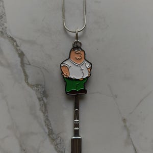May include: A silver necklace with a pendant depicting a cartoon character in a white shirt and green trousers, with a spoon-shaped design. The pendant hangs from a thin silver chain, offering a quirky and fun accessory.