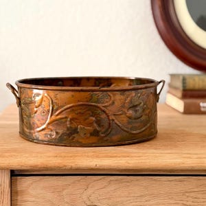 May include: A round copper basin featuring an embossed floral vine pattern along its exterior walls. The vessel has two small, integrated handles on opposite sides and displays a weathered, multi-toned patina with dark brown and green oxidation.