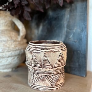 May include: A handcrafted, beige ceramic pot with a textured surface and geometric patterns. The pot features a cylindrical shape with a stacked design, showcasing triangular and linear details. The interior is a darker brown, and the pot sits on a wooden surface.