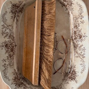 May include: An oval ceramic platter with a brown floral pattern and a crackled glaze finish. Resting on the platter is a wooden hand brush with coarse, light brown bristles and a pair of thin, round-framed glasses with brown temple tips.