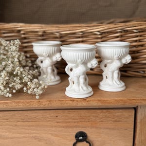 May include: Three white ceramic egg cups, each featuring a sculpted cherub figure supporting a fluted bowl. The cherubs are depicted in a standing position, holding the circular, ribbed containers above their heads on a circular base.