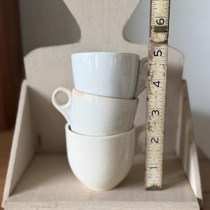 May include: Three stacked ceramic cups with a light cream glaze and visible crackle finish. A vintage wooden folding ruler is placed vertically next to the cups, showing a total height of approximately 6.5 inches for the stack.