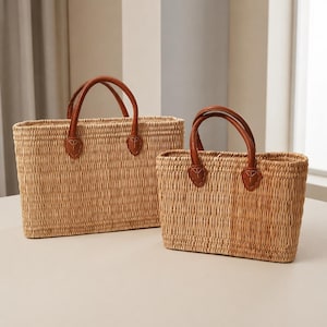 Puede incluir: Dos bolsas de mano rectangulares de paja tejida con asas de cuero marrón. Las asas están unidas a las bolsas con detalles de cuero marrón. Las bolsas son de diferentes tamaños y están sobre una superficie clara.