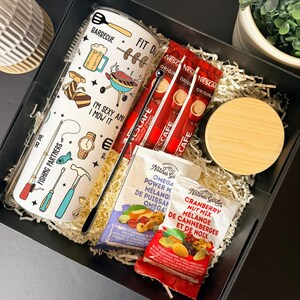 Puede incluir: Una caja de regalo con un vaso blanco con ilustraciones temáticas de barbacoa y pesca y texto. La caja también contiene paquetes de café, paquetes de mezcla de nueces y un posavasos de madera, todo en papel triturado.