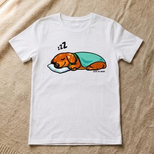 以下が含まれることがあります： 白い半袖Tシャツ。白い枕の上でティール色の毛布をかけて眠る茶色のダックスフントのグラフィックが描かれています。イラストには犬の上に黒い「zzz」の文字があり、犬の下には「COCOA THE WIENER」という文字がプリントされています。
