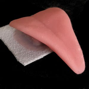 Könnte beinhalten: Eine rosa Zungenprothese aus Silikon wird auf einer weißen, strukturierten Oberfläche gezeigt.