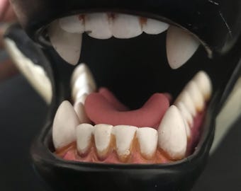 May include: Close-up of a black plastic animal mouth with white teeth and a pink tongue. The mouth is open wide, showing the teeth and tongue.