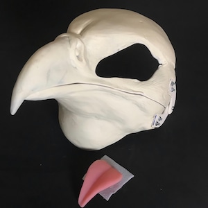 May include: A white unfinished bird mask with a separate pink tongue. The mask has a large beak and a hole for the eyes. The mask is made of a smooth material and has a textured surface.