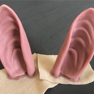 Puede incluir: Dos pares de orejas puntiagudas de silicona de color carne. Las orejas están sobre una superficie de tela beige.