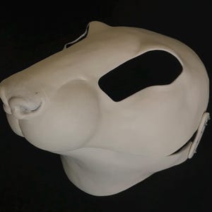 Puede incluir: Una máscara de animal de piel blanca sin terminar con agujeros negros para los ojos y una nariz. La máscara está hecha de espuma y tiene una correa en la parte posterior.