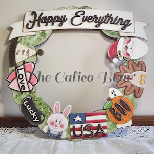 Könnte beinhalten: Ein Holz-Adventskranz mit den Worten "Happy Everything" auf einem Banner. Der Kranz ist mit verschiedenen festlichen Bildern verziert, darunter ein Kaninchen, ein Kürbis, ein Truthahn, ein Weihnachtsmann, ein Herz mit dem Wort "Love", ein Hut mit den Worten "Happy New Year" und ein Schild mit der Aufschrift "USA".