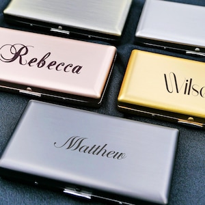 Può includere: Una collezione di porta biglietti da visita personalizzati in metallo in vari colori, tra cui argento, oro e oro rosa. Ogni porta biglietti è inciso con un nome, come "Rebecca" o "Wilson", in un elegante carattere. I porta biglietti sono rettangolari con chiusura a cerniera.