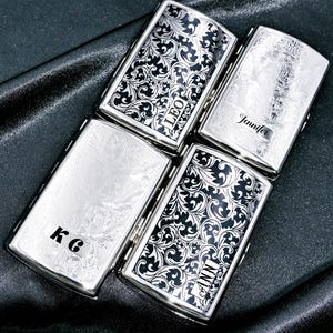 May include: Four silver cigarette cases with engraved details. Two cases display black and white floral patterns, the others have engraved designs. Each case is personalised with a name or initials: "LEO", "Jennifer", "KG", and "ANN".