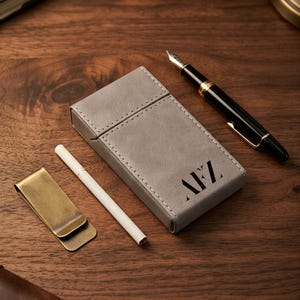 May include: A rectangular grey cigarette case with white stitching and the black monogram letters AFZ printed on the front. Beside the case lies a single white cigarette, a brass-toned metal money clip, and a black fountain pen with gold trim.