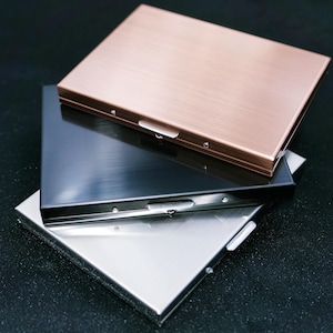 May include: Three metal card cases in copper, black, and silver, stacked. Each case has a rectangular shape with a brushed metal finish and a small latch. The cases are on a dark background.