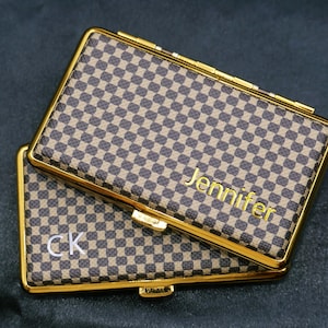 Puede incluir: Dos estuches rectangulares con ribete dorado y un patrón de cuadros marrones y beige. Un estuche tiene el nombre "Jennifer" en letras doradas, y el otro tiene las iniciales "CK" en letras blancas. Los estuches están apilados.