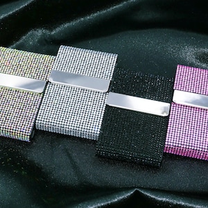 Puede incluir: Cuatro estuches rectangulares cubiertos de pequeños y brillantes pedrería. Los estuches son en tonos plateado, negro y rosa, cada uno con una barra de metal plateado en la parte delantera. Probablemente son tarjeteros o accesorios similares.