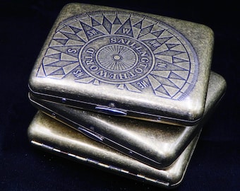 May include: Three stacked brass cigarette cases with a compass design and the text "Sailing to the World" on the top case.