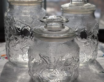 May include: Three clear glass jars with lids. The jars have a textured surface with a floral design. The jars are of varying sizes and are arranged on a white cloth with embroidered flowers.