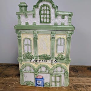 May include: A ceramic cookie jar shaped like a building with a pale yellow facade, green trim, and a white roof. The front features the text "POST OFFICE" above a blue mailbox with the word "MAIL" printed on it, along with arched windows.