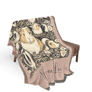 Peut inclure: Un plaid tissé présentant un motif répétitif de cochons d'Inde et de marguerites blanches sur fond sombre. La partie inférieure est d'une couleur rose pâle unie avec le texte manuscrit "Squeak Love" en écriture noire et des bords à franges.