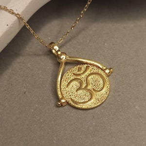 May include: A gold-colored necklace featuring a pendant with an Om symbol. The pendant is round with a textured surface and is suspended from a delicate chain. The necklace is displayed on a neutral gray surface.