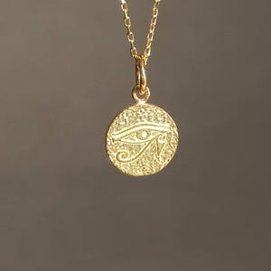 May include: A gold-colored necklace featuring a circular pendant with a textured surface. The pendant displays the Eye of Horus symbol, a stylized eye with intricate details. The chain is delicate and complements the pendant's design.
