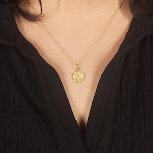 May include: A gold-colored pendant necklace with a circular charm. The charm has an intricate design and is suspended from a delicate chain. The necklace is worn against a black top.